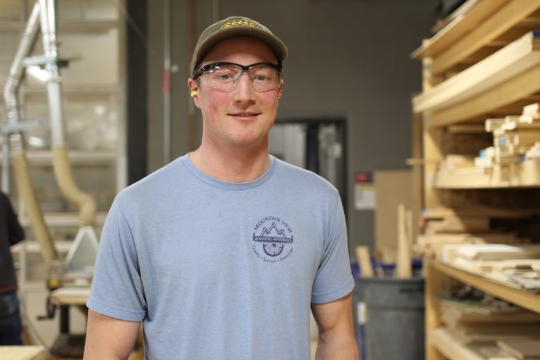 A man wearing a ball cap, safety goggles and ear plugs stands in front of a shelf carrying planks of wood inside a carpentry lab.