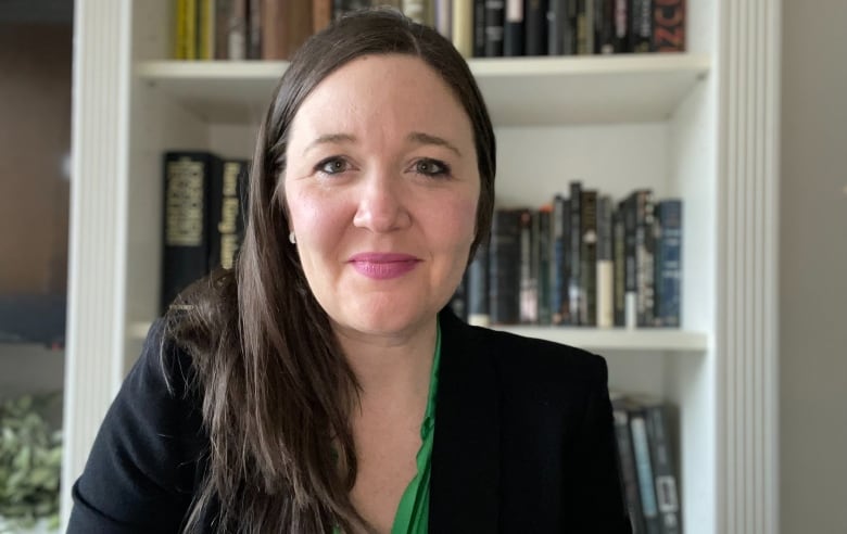 A woman with long brown hair, wearing a black blazer and green blouse, sits in front of a bookshelf.