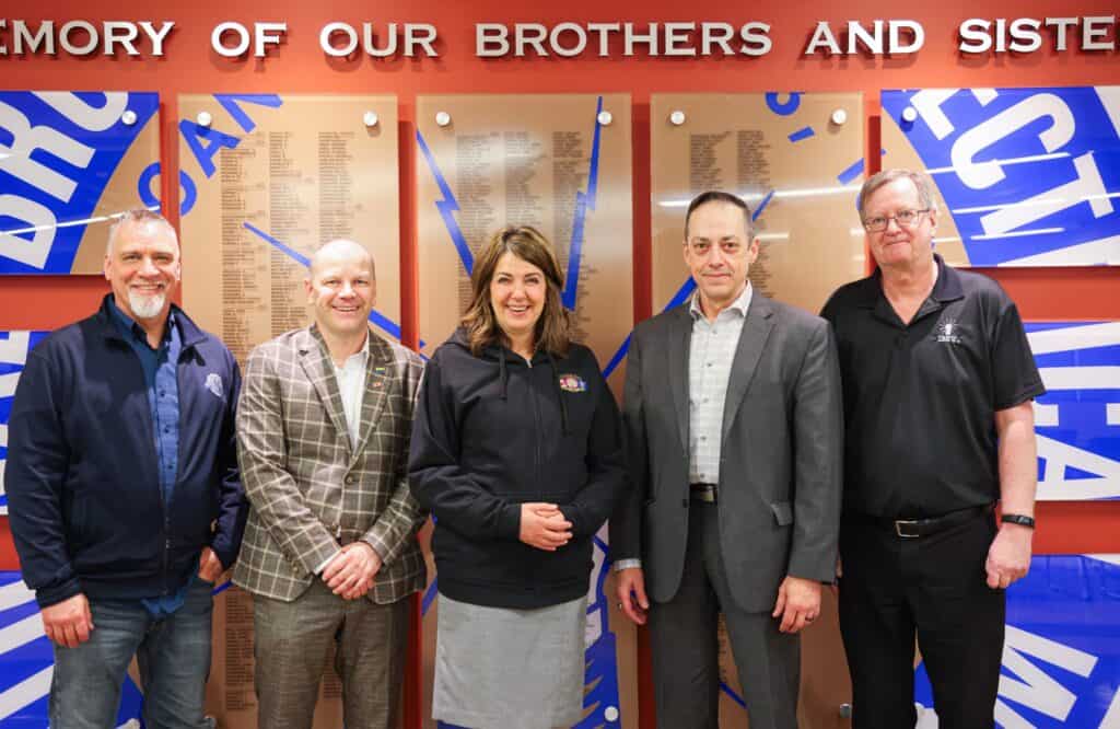 Members of the IBEW Local 424 leadership team (Todd Chrunik (Training Director, IBEW 424), Scott Crichton, Michael Reinhart, Bill Begemann (International Representative, IBEW Canada) are joined by Alberta Premier, Danielle Smith (centre). They stand in front of the IBEW 424 Tribute Wall, honouring members who have passed away.