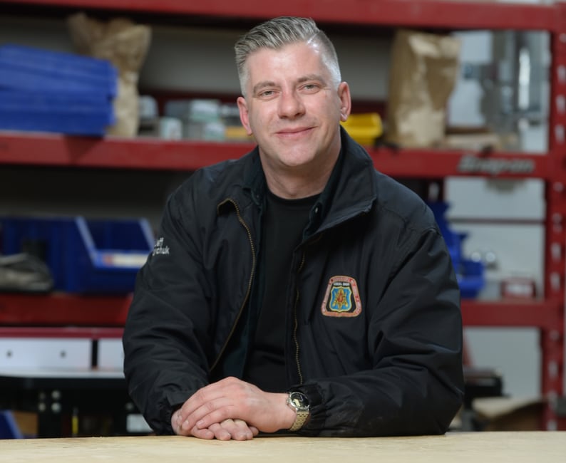 A smiling man in a black zip-up jacket leans against a table indoors, with shelving storing construction equipment seen behind him.