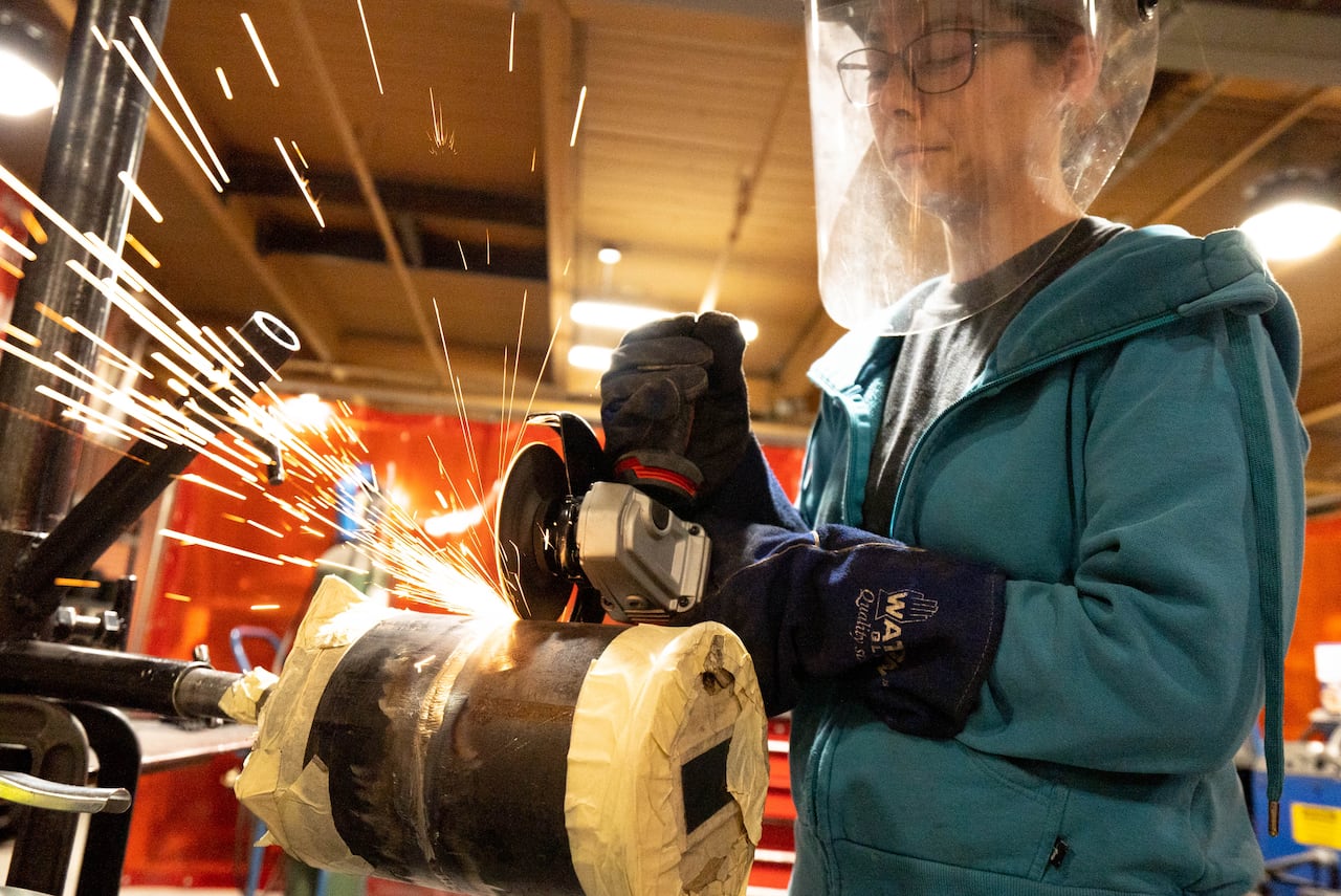 A woman in protective gear, including a face shield and gloves, works at a metal grinder station in a training facility. 